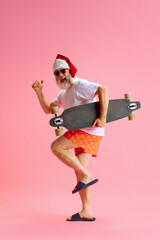 Happy funny nice mature man in sunglasses and Santa Claus festive hat having fun holding skate board against pastel pink studio background.