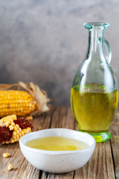 Corn oil in ceramic bowl and in jug, corn cobs over rustic wooden table with grey background