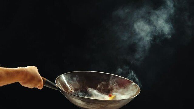 Man cooking delicious wok noodles, frying vegetables in wok pan, preparing ingredients against black background. Concept of Asian food, cuisine, restaurant, taste, cooking, recipe