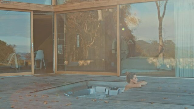 Woman unwinds in a jacuzzi on a wooden deck, framed by a modern cabin.