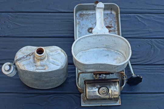 an empty aluminum old camping mug with a handle stands on an iron industrial retro rusty camping primus for heating water along with an aluminum open army gray flask stands on a wooden black table