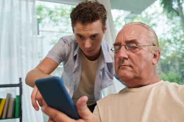 Naklejka na ściany i meble Young man helping grandfather to install dating application on smartphone