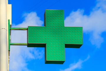 Green medical cross indicating the location of a pharmacy on a city street. Banner medical cross on a building against a background of blue sky and clouds.Pharmacy sign.