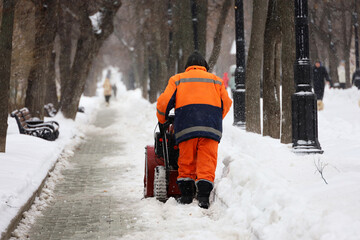 Communal services worker in uniform with a snowplow clears snow on a sidewalk. Man during snow...