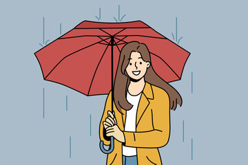 Happy woman with umbrella walks in rain along autumn street and looks at screen with wide smile. Cheerful girl dressed in casual clothes uses umbrella to avoid getting wet and catching cold.
