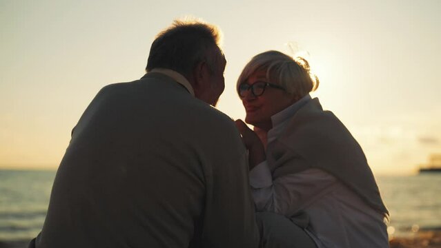 Romantic senior couple sitting on sea beach talking resting together. Man kissing woman hands at sunset in evening. Family laughing spending time together. Romantic relationship, lifelong marriage.