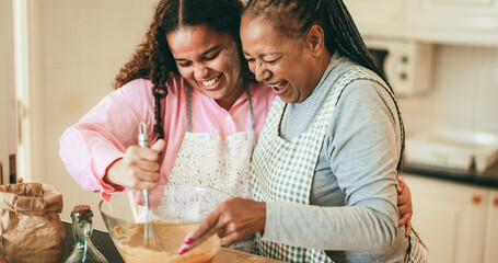 African mother and daughter having fun preparing fruit cake at home - Winter holidays and family concept - Soft focus on senior woman mouth - obrazy, fototapety, plakaty