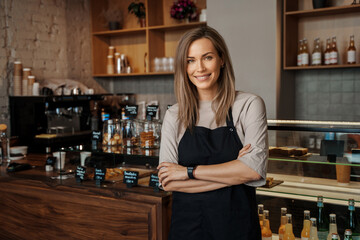 Smiling woman in apron standing confidently in a cozy cafe environment, with shelves of flowers in the background