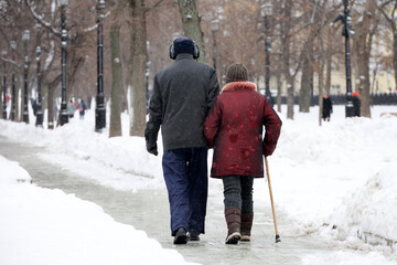 Elderly woman witj cane and man walking in winter park, rear view. Old couple in warm clothes...