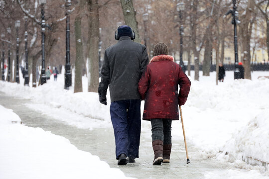 Elderly woman witj cane and man walking in winter park, rear view. Old couple in warm clothes during snow weather, concept of old age