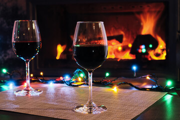 Two red wine glasses and lights decoration on burning fireplace background. Wine by the fireplace.