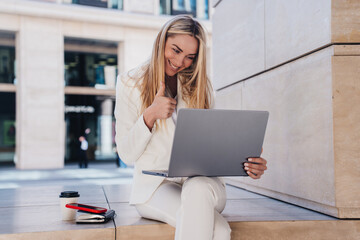 Grateful young entrepreneur in white business suit sitting outside makes video call using laptop shows thumb up toothy smiles. Cheerful blonde student girl passed exam shares great news with parents.
