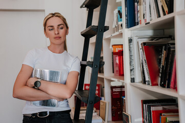 Thoughtful young blonde lady standing at book store looks aside with sad face expression holds...