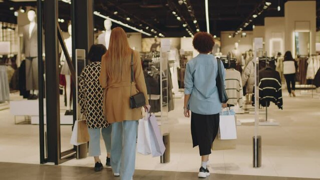 Full length back view shot of three multiethnic young women entering stylish clothes store in big modern shopping mall