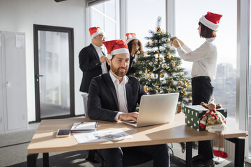 Attractive Caucasian businessman employee in suit and santa hat sitting at desk using laptop in christmas decorated office. Young corporate worker in festive workplace looking at screen of computer.