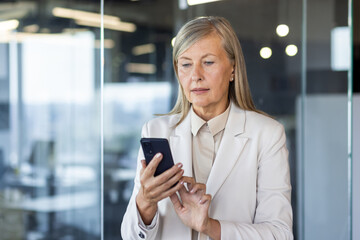 Fototapeta premium Serious gray-haired businesswoman standing in the office and using the phone, typing and reading messages, making calls. Close-up photo.