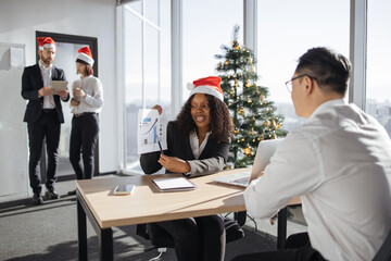 Christmas time. Smiling African businesswoman in jacket and Santa hat showing documents with graphs and charts to asian employee while coworkers decorating office and Christmas tree.