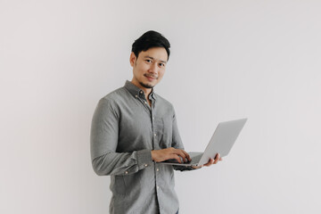 Asian man with beard wear grey shirt typing keyboard notebook and holding laptop with happy smiling face, working and looking at camera, standing isolated over white background wall.