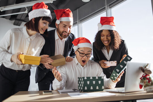 Secret Santa. Smiling Asian businessman in Santa hat unpacks a Christmas present from multinationals colleagues at work standing around him. New Year's eve.