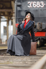 A beautiful girl in a black coat and hat with a suitcase in her hands near an old steam locomotive....