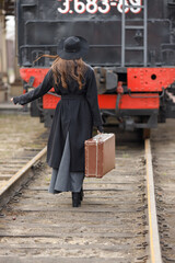 A beautiful girl in a black coat and hat with a suitcase in her hands near an old steam locomotive. A young woman with long dark hair. Vintage portrait of the last century, retro journey.