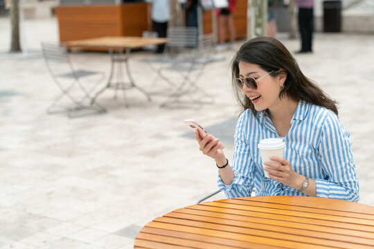 happy asian taiwanese woman executive holding coffee and reading good news about business success on phone at outdoor seating on urban background.