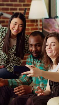 Vertical video BIPOC friends taking group pictures with selfie camera smartphone at apartment party. Joyful mates in modern home posing for photo with wine and champagne glasses, making gestures