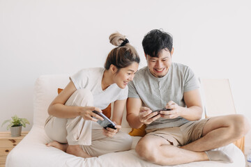 Asian Thai couple using mobile phone together while sitting on white sofa in apartment room, both woman and man happy and enjoy playing game online, family spending time together.