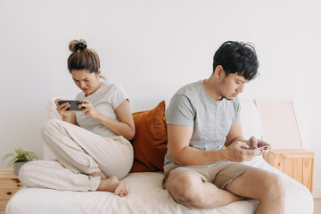 Asian Thai couple sitting back to back on sofa at home, Both man and woman using mobile phone, playing game online, ignoring living in apartment room.