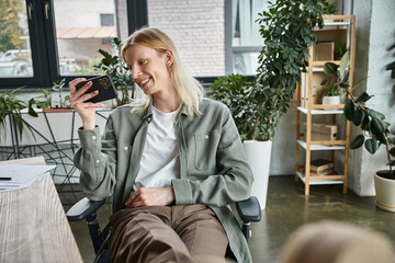 joyful young non binary person with long hair smiling at mobile phone while at office, business