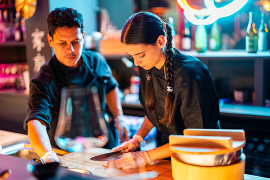 Young female chef standing while cutting meat with knife on chopping board while working with male coworker at illuminated sushi bar