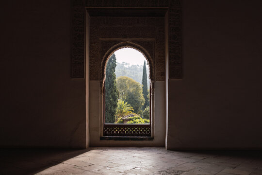 Lush garden seen through arached window in dark room of historic Alhambra fortress in Granada, Spain