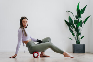 Gorgeous brunette European girl in sportswear makes exercise at home adjusts massaging roll by...