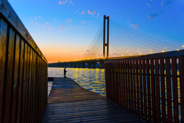 Obraz premium Wooden Dock and Fisher with a Cable-Stayed Bridge at Sunset