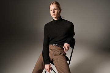 attractive non binary model in black turtleneck with ponytail sitting on black chair, fashion