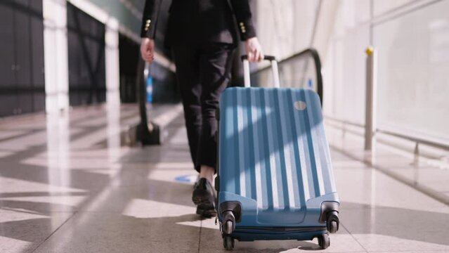 Caucasian young woman in professional attire walks with blue suitcase on an airport moving walkway, epitomizing modern business travel and active lifestyle. Female tourist with luggage move to gate