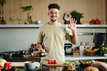 Medium shot of smiling young middle eastern chef happily waving at camera holding pan while posing at kitchen, copy space