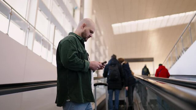 Smiling bald male traveler in casual attire stands on airport escalator, catching up on messages or planning his itinerary. Young man using online banking application for send money with mobile phone
