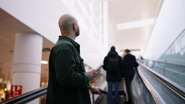 Bald Caucasian man in a green shirt is focused on his smartphone while standing on an airport escalator,  modern traveler's need to stay connected on the move while connecting to wireless via app
