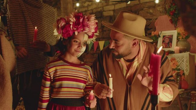 Medium shot of Latin man speaking to little daughter with half painted skeleton face while standing with other family members in backyard at night holding candles and doing ritual on Dia de Muertos