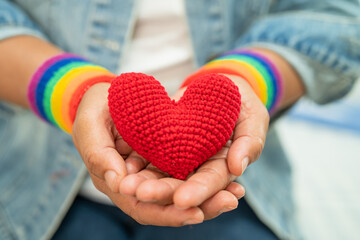 Asian lady wearing rainbow flag wristbands and hold red heart, symbol of LGBT pride month celebrate...