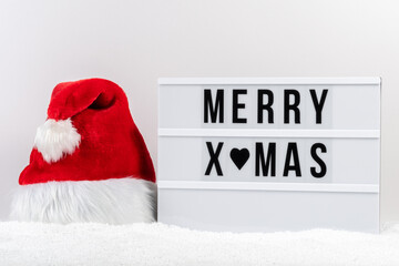 White lightbox with text MERRY XMAS and Santa Claus hat on a table with snow. Minimal Christmas or New Year concept. - Powered by Adobe