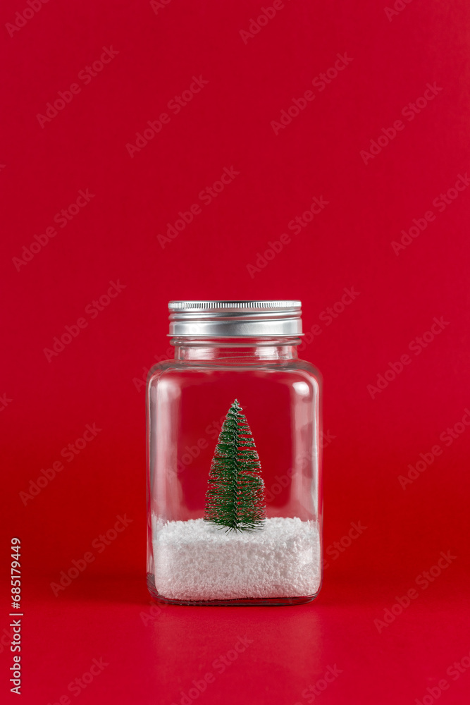 Wall mural Green Christmas tree with snow in a glass jar on red background. Minimal Christmas or New Year holiday concept.