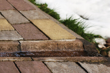 Concrete Gutter for Rainwater or Melt Water Drainage. Water from Melted Snow flowing on Concrete Gutter into Drainage System. Concrete Paver Gutter from House. - obrazy, fototapety, plakaty