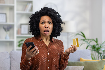 Close-up photo of African-American woman sitting on sofa at home, holding phone and credit card, looking at camera worried and shocked