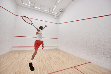 young athletic african american player with racquet jumping and playing squash in court, back view