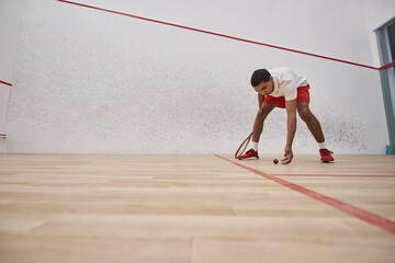 african american man in red shorts holding racquet and picking up squash ball inside of court