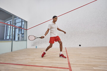 african american sportsman in red sporty shorts holding racquet while playing squash inside of court