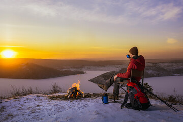 man hiker sitting near bonfire and drinking tea during camping in winter at sunset.