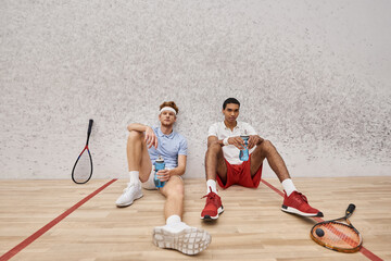 two tired interracial friends with squash racquets and bottle with water sitting on floor in court